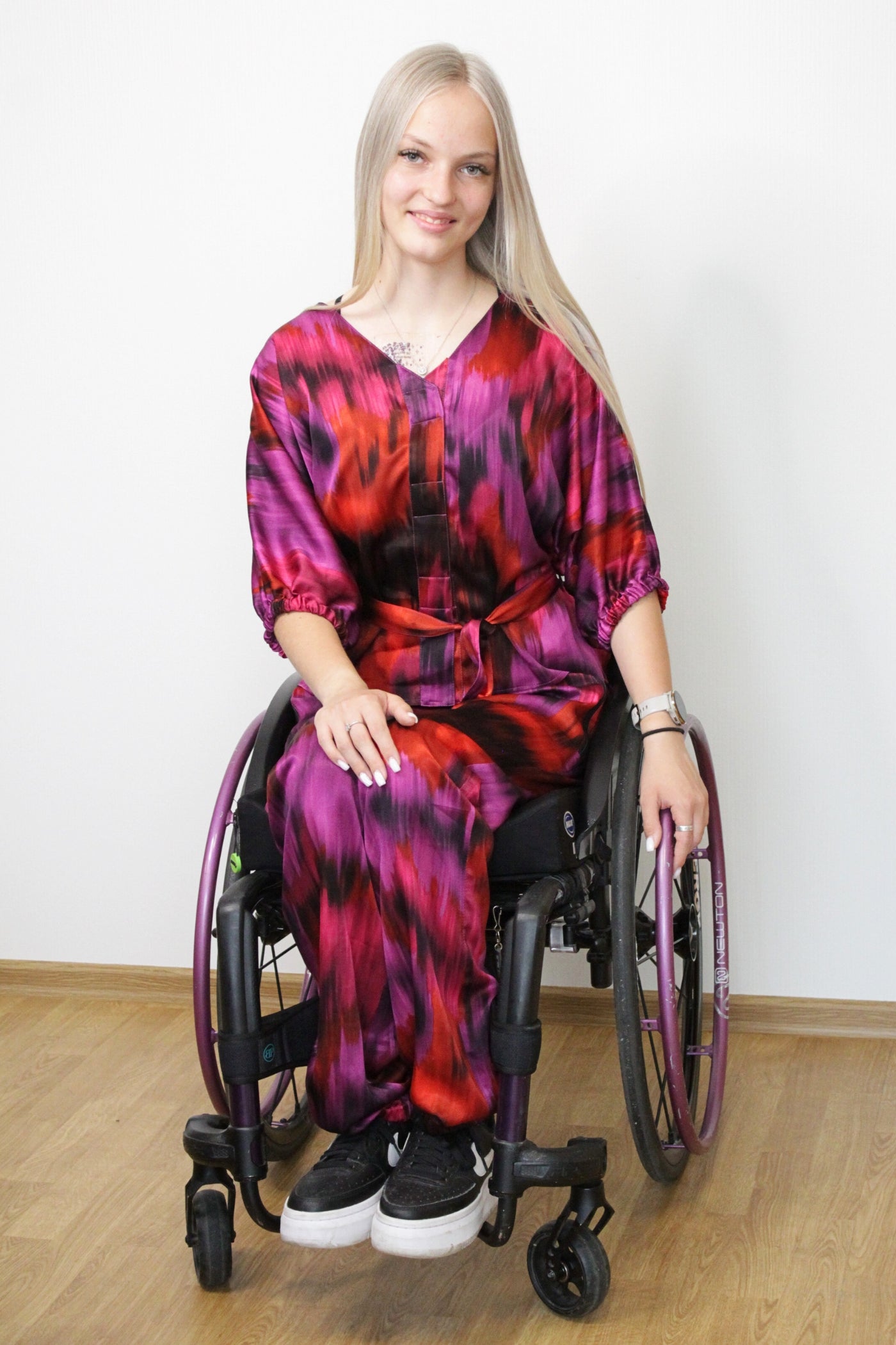Ein langes, blondes Model mit einem leichten Lächeln trägt einen luxuriösen zweiteiligen Satz aus 100 % Viskose-Satin in leuchtenden Farben wie Fuchsia, Lila und Orange. Das Set ist für Rollstuhlfahrer geeignet und verfügt über magnetische Verschlüsse, was es auch für Menschen mit eingeschränkter Handfunktion, wie Tetraplegiker, leicht an- und auszuziehen macht.