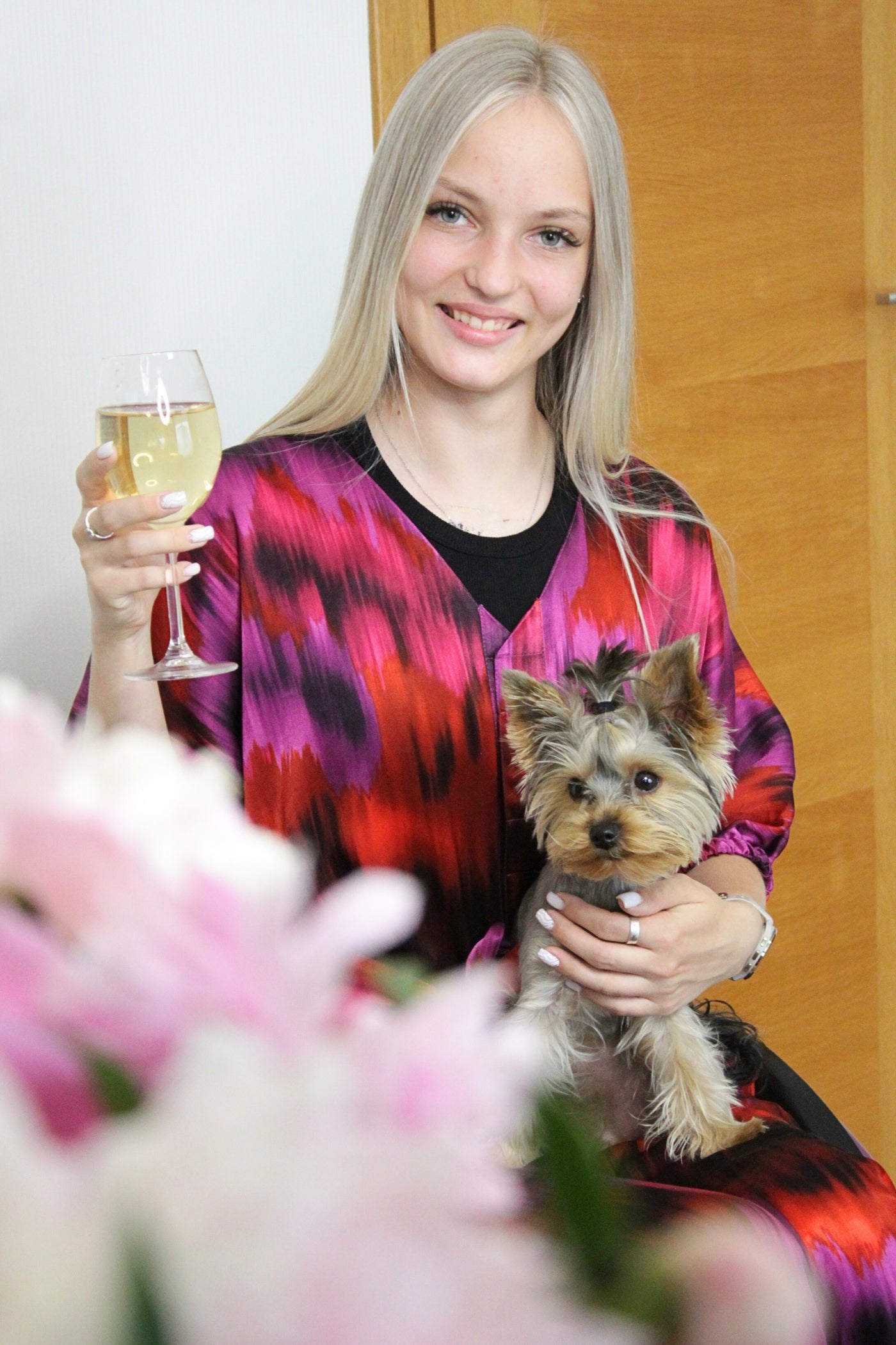 Lächelnde Frau im adaptiven Rollstuhl-Satin-Set in Pink- und Rottönen, hält ein Glas Weißwein und einen kleinen Yorkshire Terrier. Das luxuriöse zweiteilige Set ist Rollstuhl-adaptiv und für Menschen mit eingeschränkter Handfunktion geeignet.