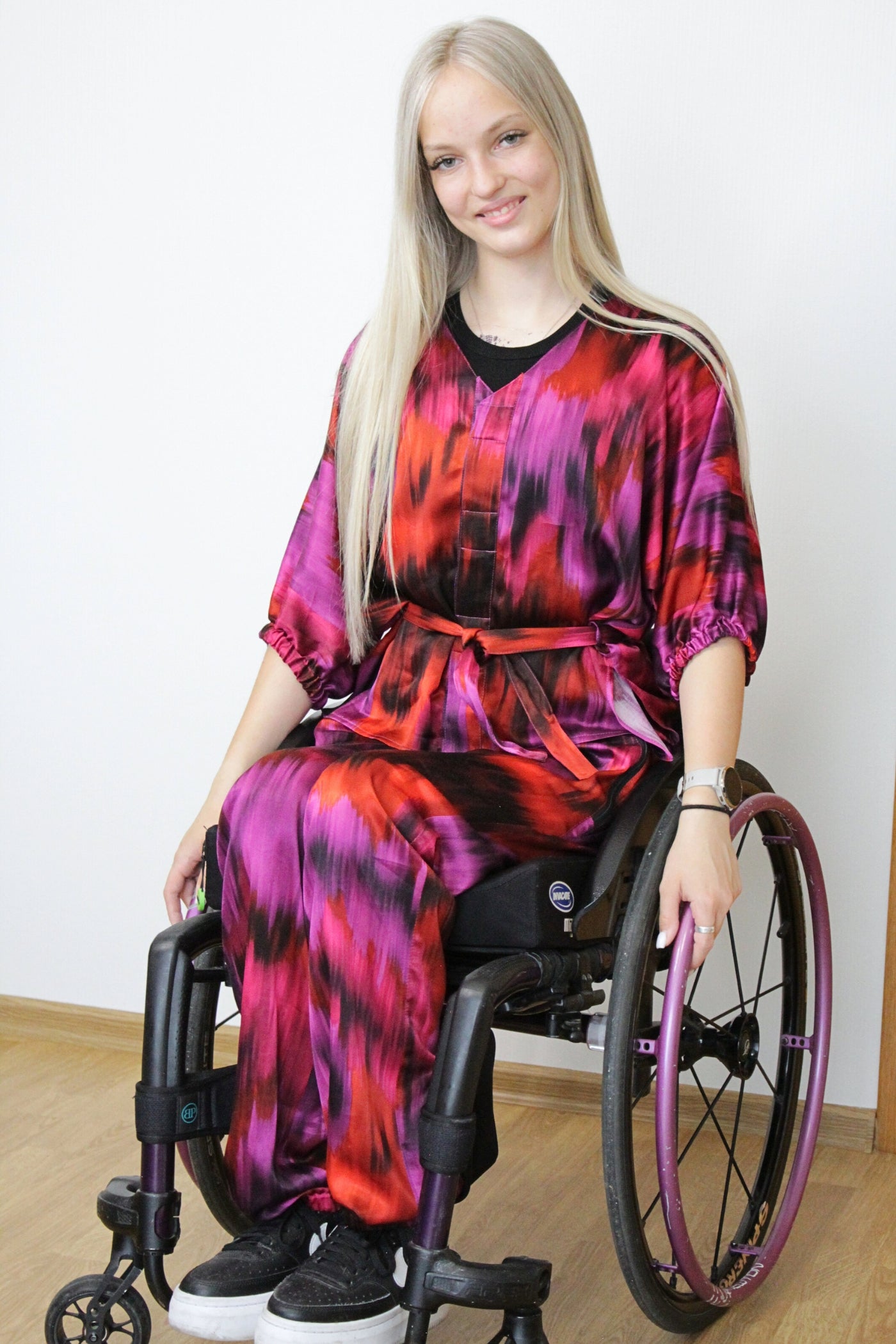 Ein langes, blondes Model mit einem leichten Lächeln trägt einen luxuriösen zweiteiligen Satz aus 100 % Viskose-Satin in leuchtenden Farben wie Fuchsia, Lila und Orange. Das Set ist für Rollstuhlfahrer geeignet und verfügt über magnetische Verschlüsse, was es auch für Menschen mit eingeschränkter Handfunktion, wie Tetraplegiker, leicht an- und auszuziehen macht. Das Model trägt außerdem eine Armbanduhr und mehrere Ringe.