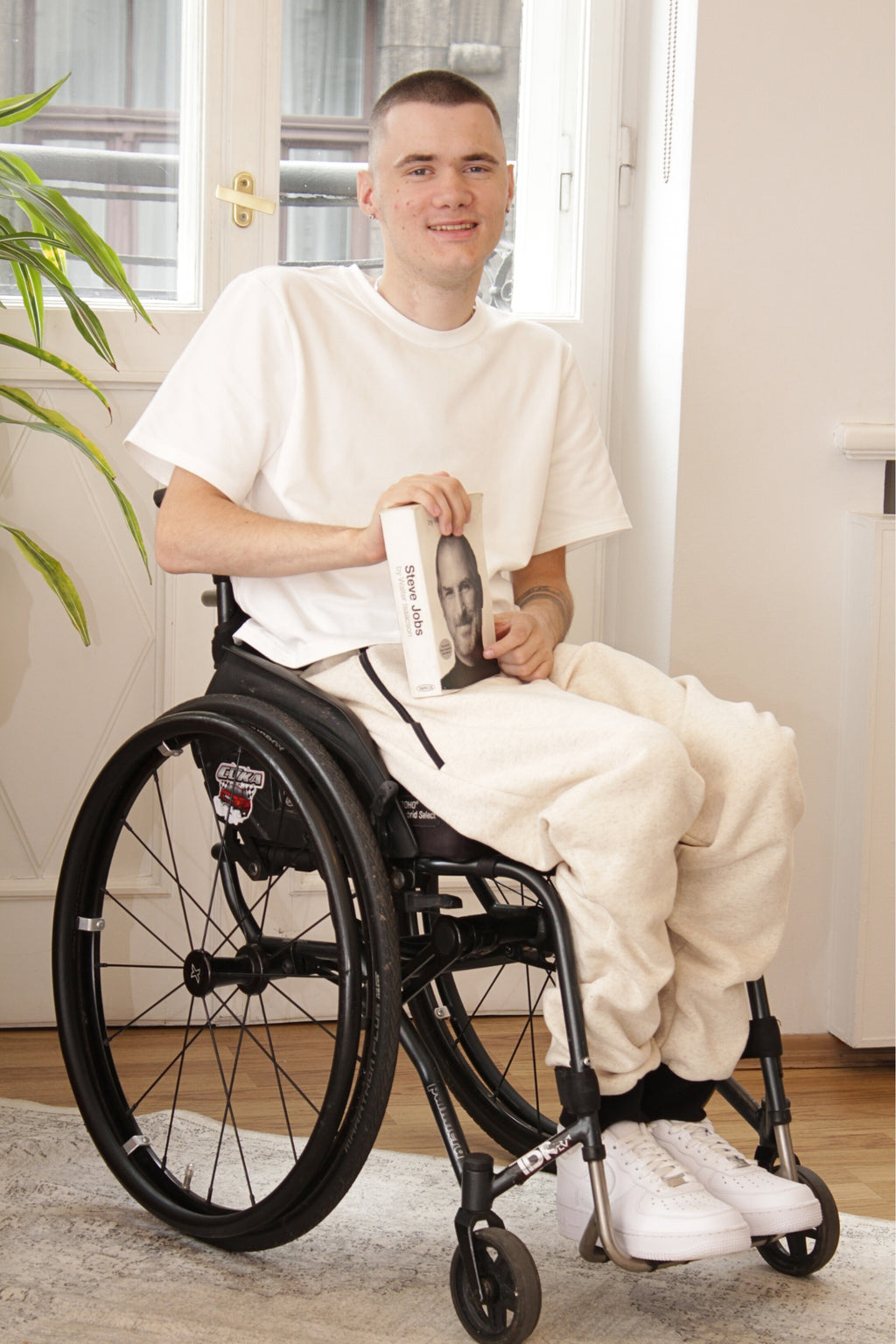 Ein lächelnder junger Mann mit kurz geschnittenen Haaren sitzt in einem Rollstuhl und hält ein Buch mit dem Titel „Steve Jobs“ in der Hand. Er trägt adaptive Kleidung, bestehend aus einem weißen T-Shirt und einer hellbeigen Jogginghose. Seine weißen Turnschuhe sind deutlich sichtbar. Im Hintergrund ist ein Fenster zu sehen und eine Pflanze lugt vom linken Bildrand herein.