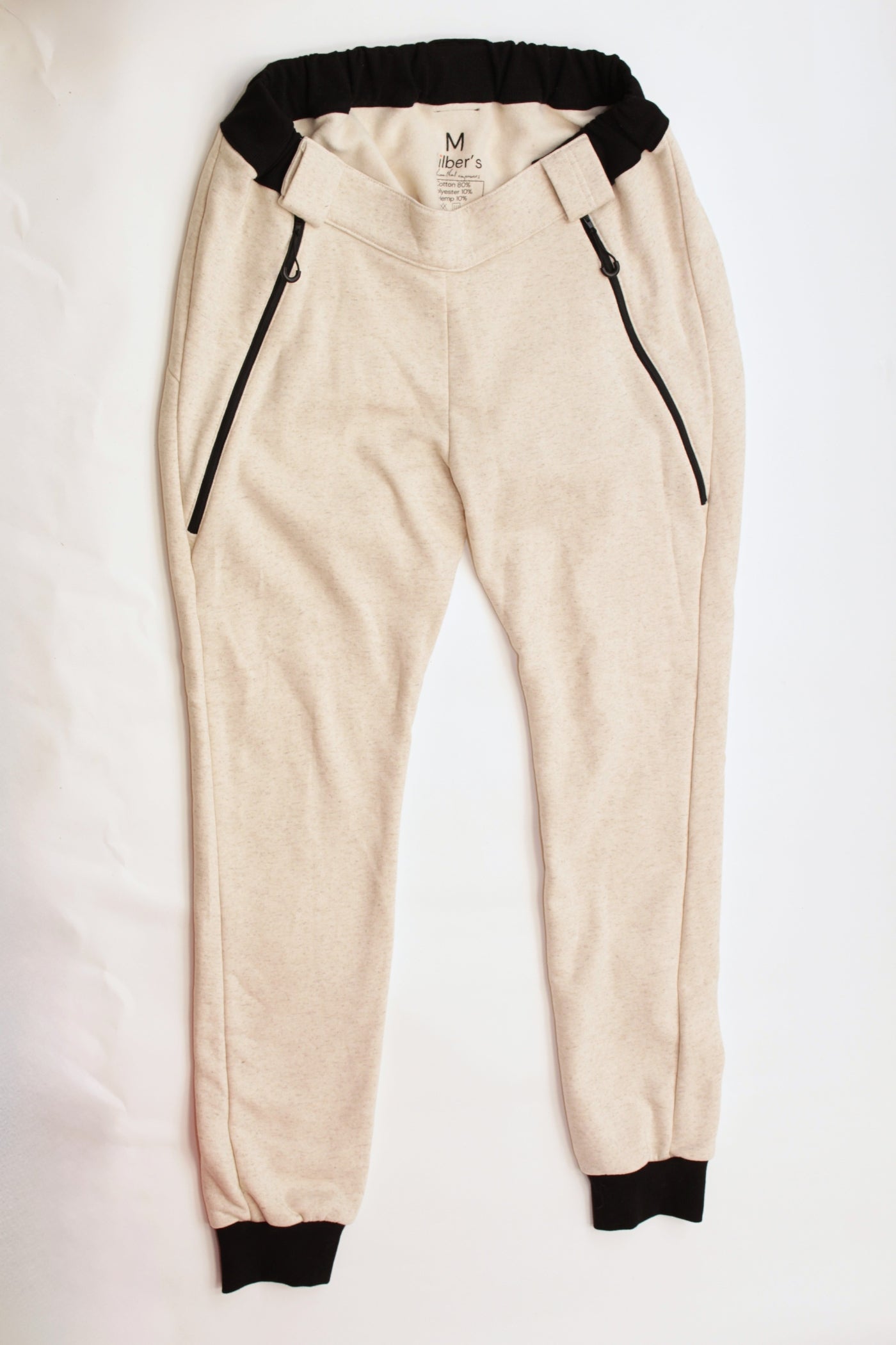 Here is the alt text you requested:

Eine beigefarbene Jogginghose mit schwarzen Bündchen an Bund und Knöcheln sowie schwarzen Reißverschlusstaschen, flach ausgelegt auf einem weißen Hintergrund. Es ist eine adaptive Rollstuhlhose von Vilber's, die speziell für Menschen mit Behinderungen entwickelt wurde.