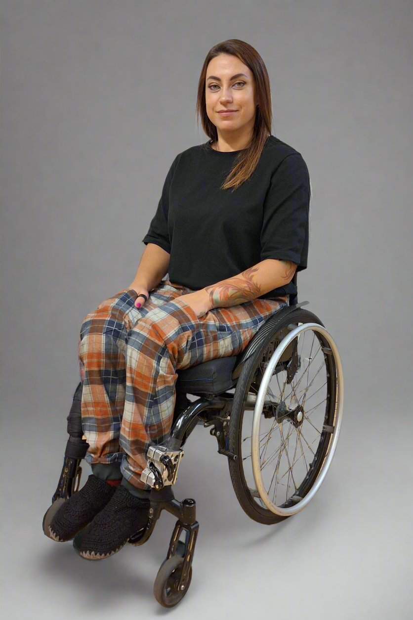 Eine Frau im Rollstuhl trägt ein schwarzes Oberteil, das an der Schulter geöffnet wird. Eine helfende Person assistiert beim Schließen des Oberteils. Dieses Bild zeigt adaptive Kleidung und Rollstuhlkleidung.