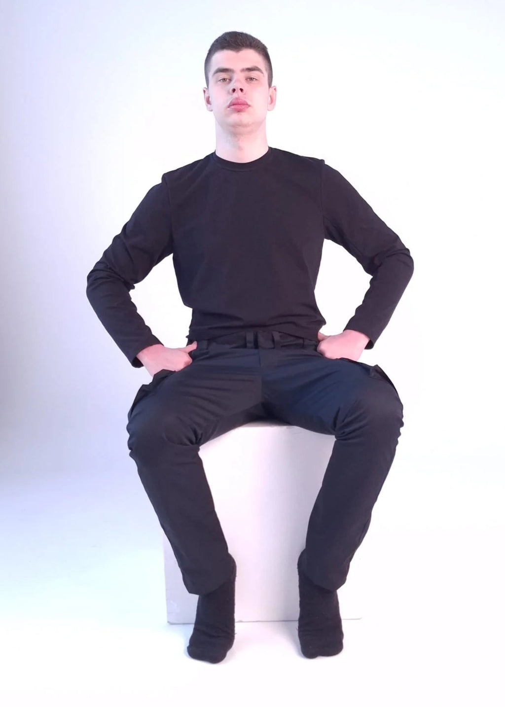 Person wearing a black wheelchair adaptive clothing sitting on a white stool against a white background