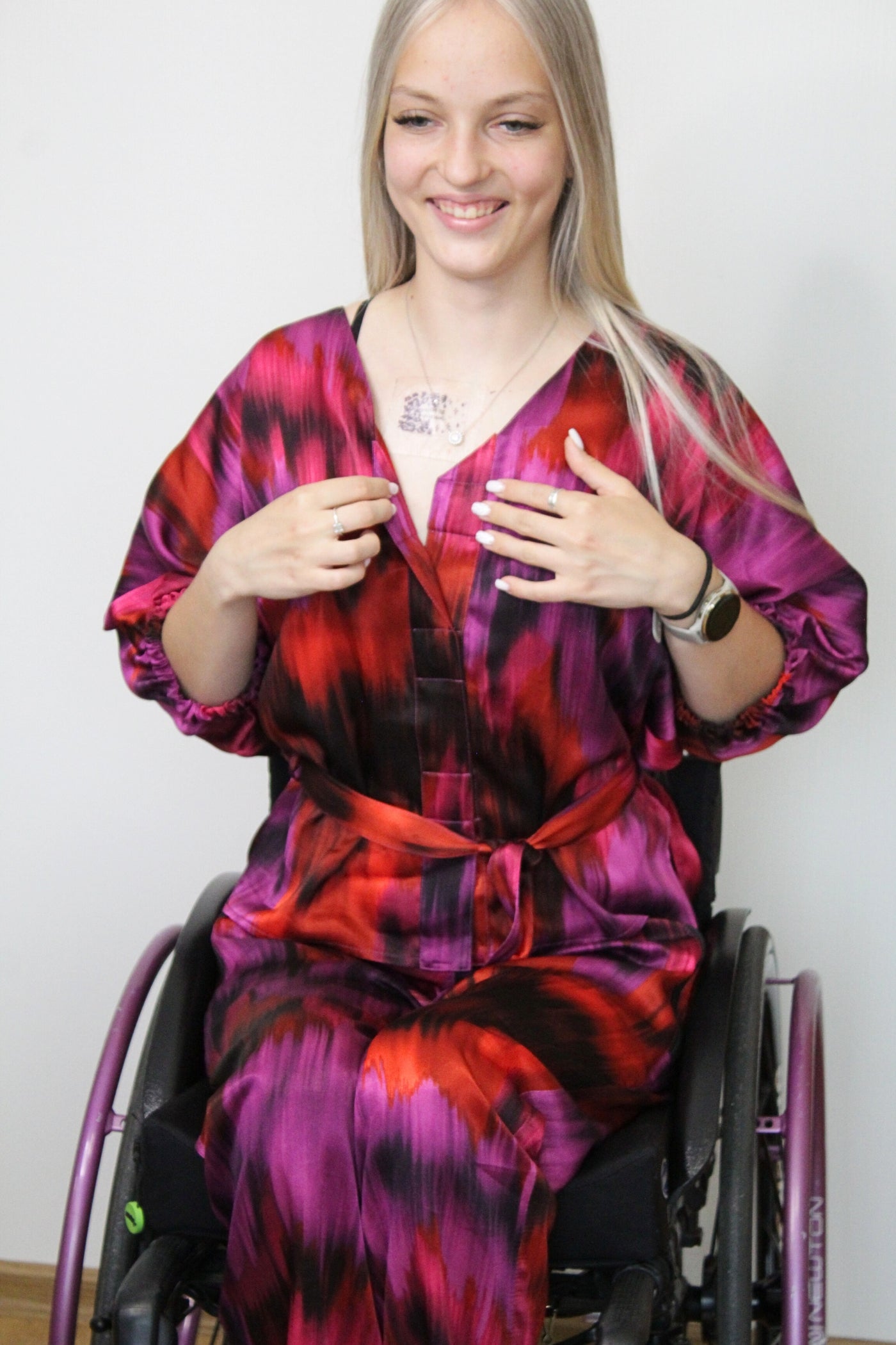 An young blonde woman who is a wheelchair model is showing the magnetic closure of premium 100% viscose satin blouse. The color of the set is fuchsia with darker shades. The set has batik pattern. The set is made for special occasions. 
